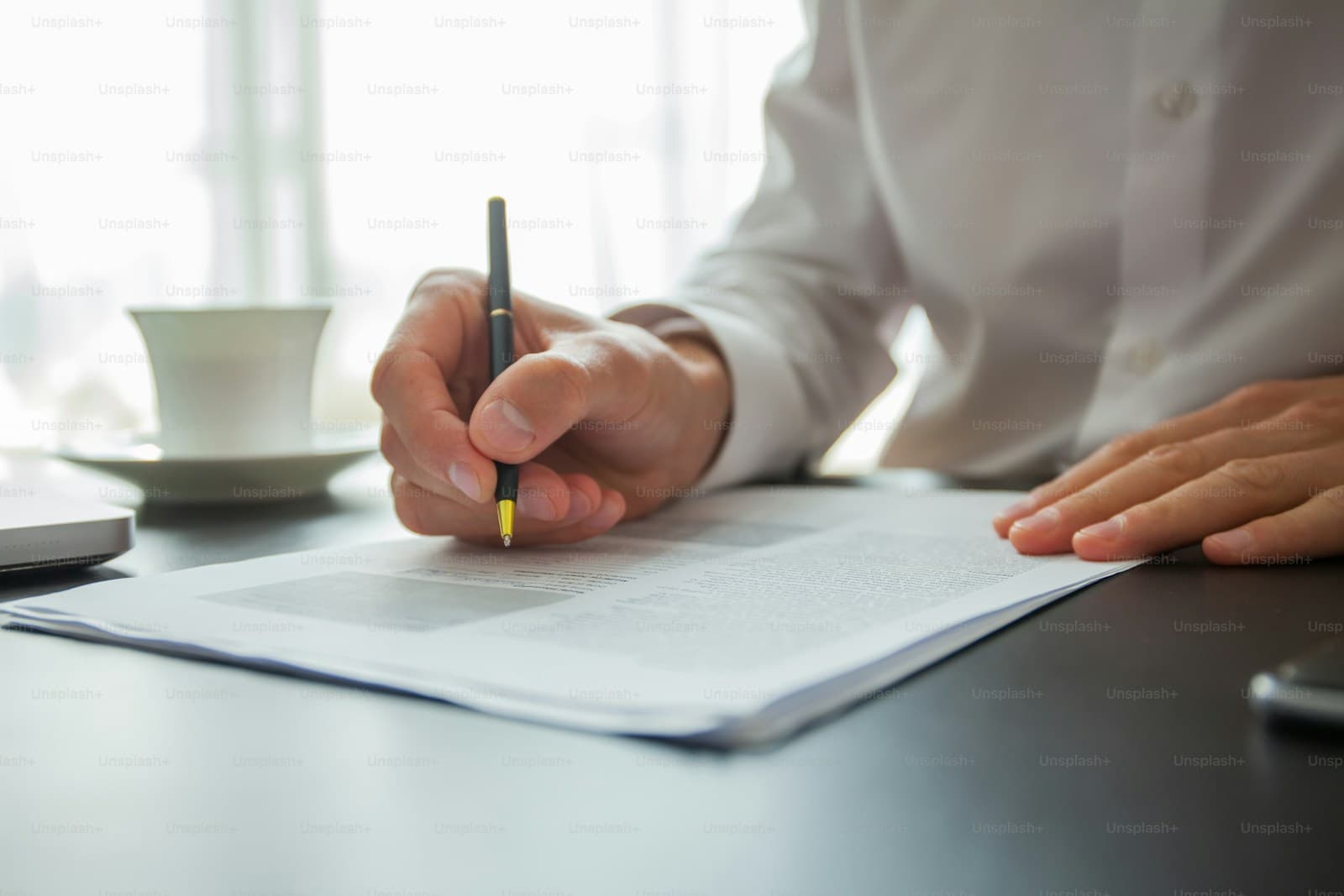Person in a white shirt writing with a black pen beside a coffee cup and saucer